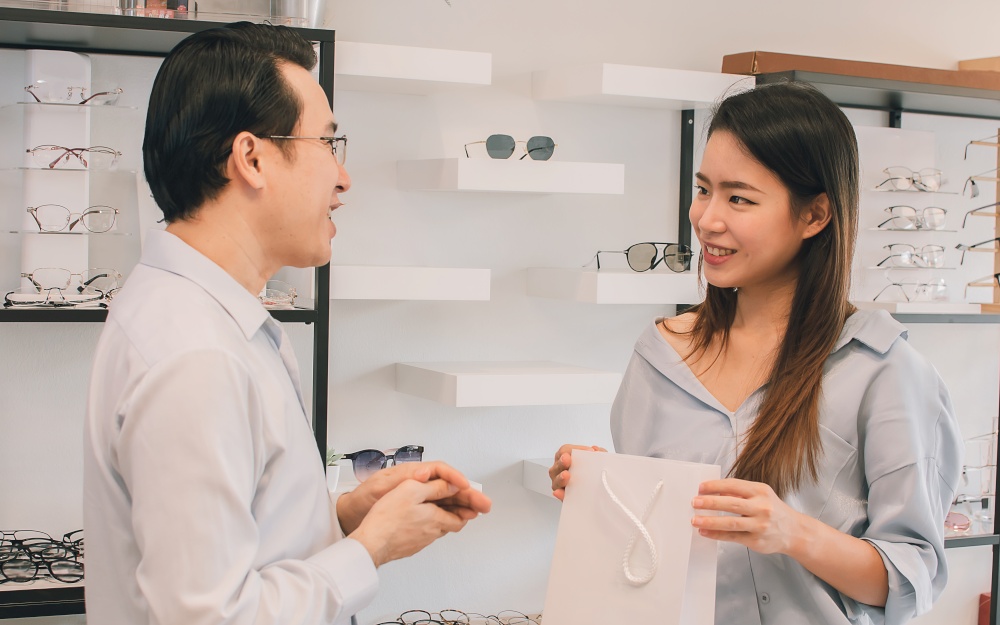 Asian male ophthalmologist give advice or consultation and selling eyeglasses to female customer at store. Optical and Sales Promotion Concept.
