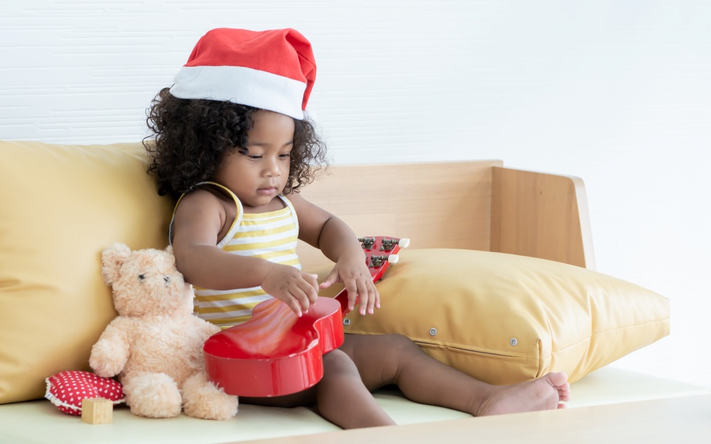 Little african black girl with afro hair wearing santa hat playing ukulele in living room at home. Christmas Celebration Concept.