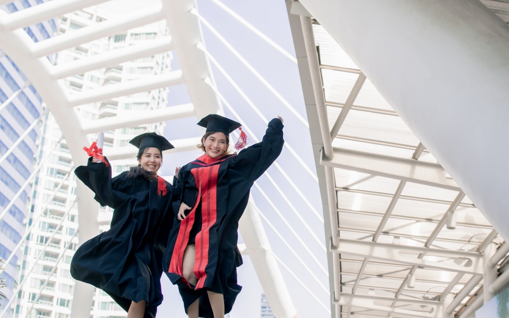 Two asian female students wearing uniform, cap, jumping and smiling with happiness after graduation. Education Concept.