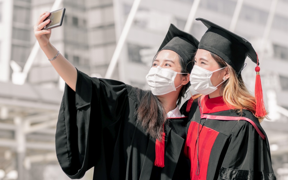 Two asian female students wearing face mask to protect virus and using mobile phone to take selfie. Graduation, Education, Technology and Health Concept.