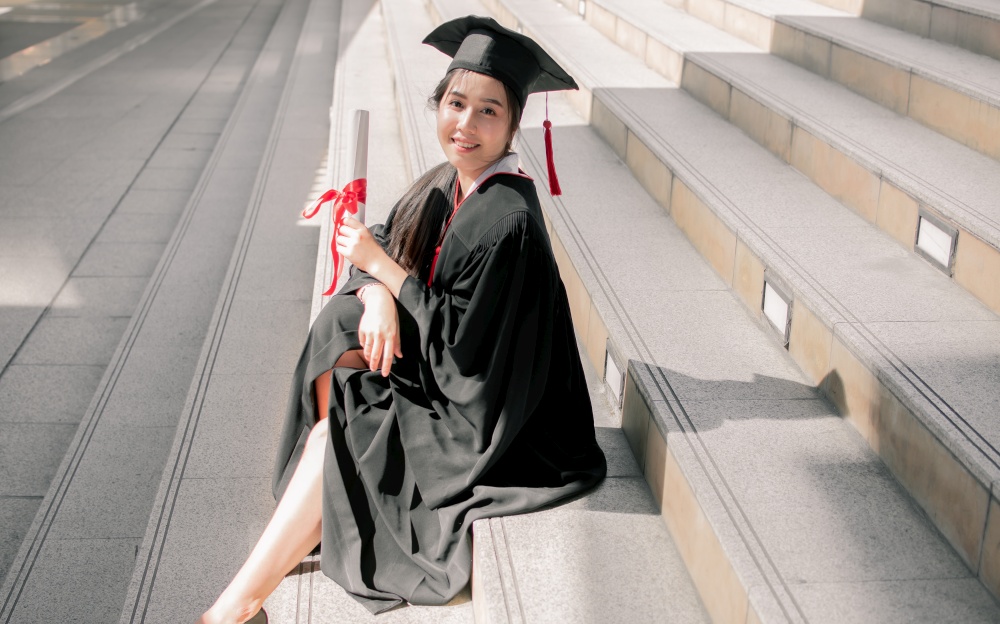 Portrait of Asian female student wearing graduated uniform and holding certification while sitting on stairs. Education Concept.