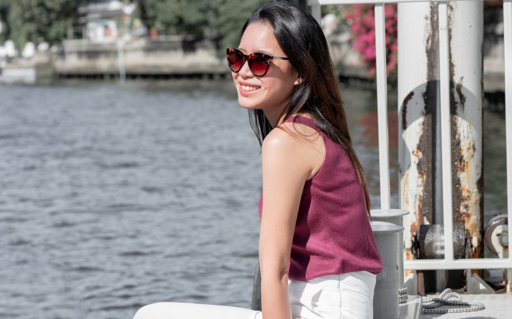 Asian beautiful woman wearing sunglasses, sitting and waiting for a boat near the river on vacation day. Summer and Travel Concept.