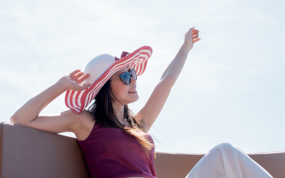 Asian beautiful woman wearing hat and sunglasses, waving her hand while sitting on boat with background of beautiful landscape. Lifestyle and Travel Concept.