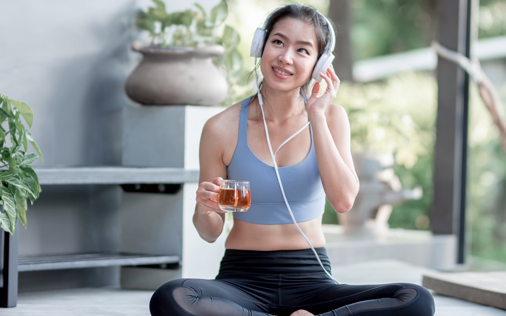 Asian beautiful and sportive woman drinking hot tea, wearing headphone  and listening to music at home in the morning. Healthy Concept.