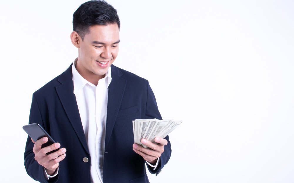 Asian business man wearing suit, using mobile phone and holding money with smiling happy face and standing on white background