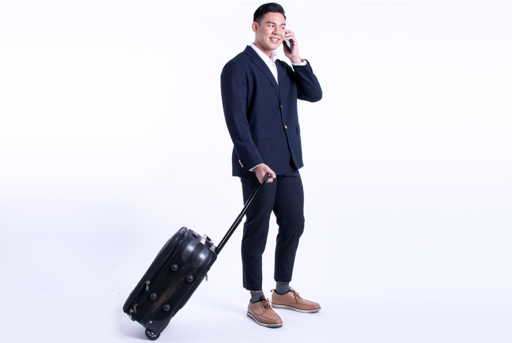 Asian business man talking on mobile phone and holding baggage for traveling, standing on white background. Technology and Vacation Concept.