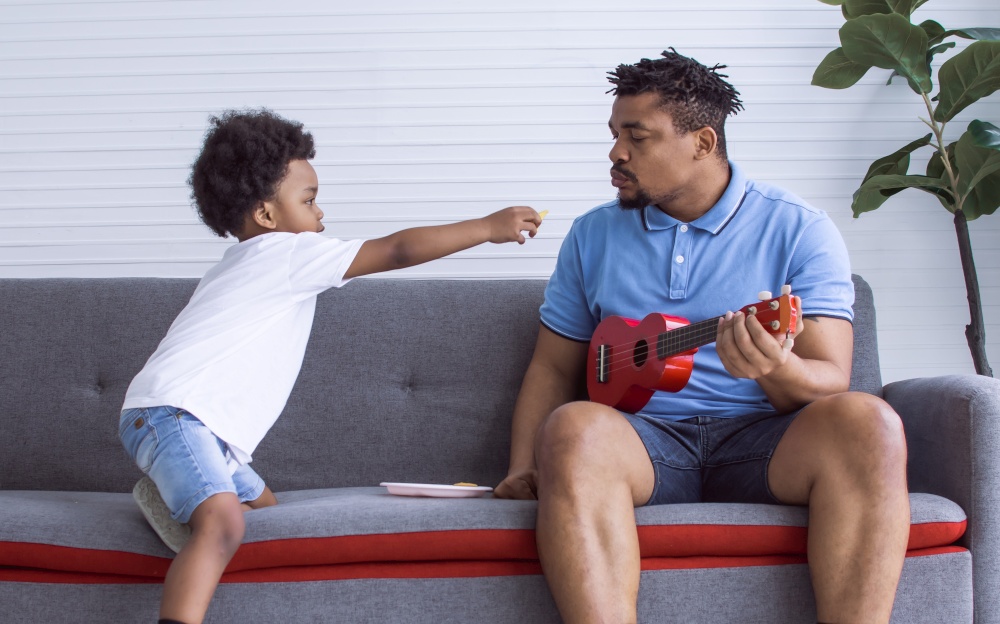 African father and little son are playing ukulele and sitting on sofa in living room at home. Family and Education Concept.