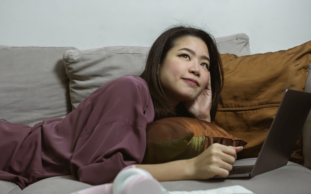 Asian young beautiful woman laying down on sofa and chatting in laptop while relaxing in living room at home. Lifestyle, Technology and New Normal Concept.