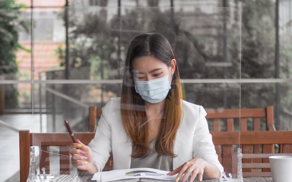 Asian business woman wearing mask, sitting behind glass partition to protect virus and working outside. Business, New Normal and Healthy concept.