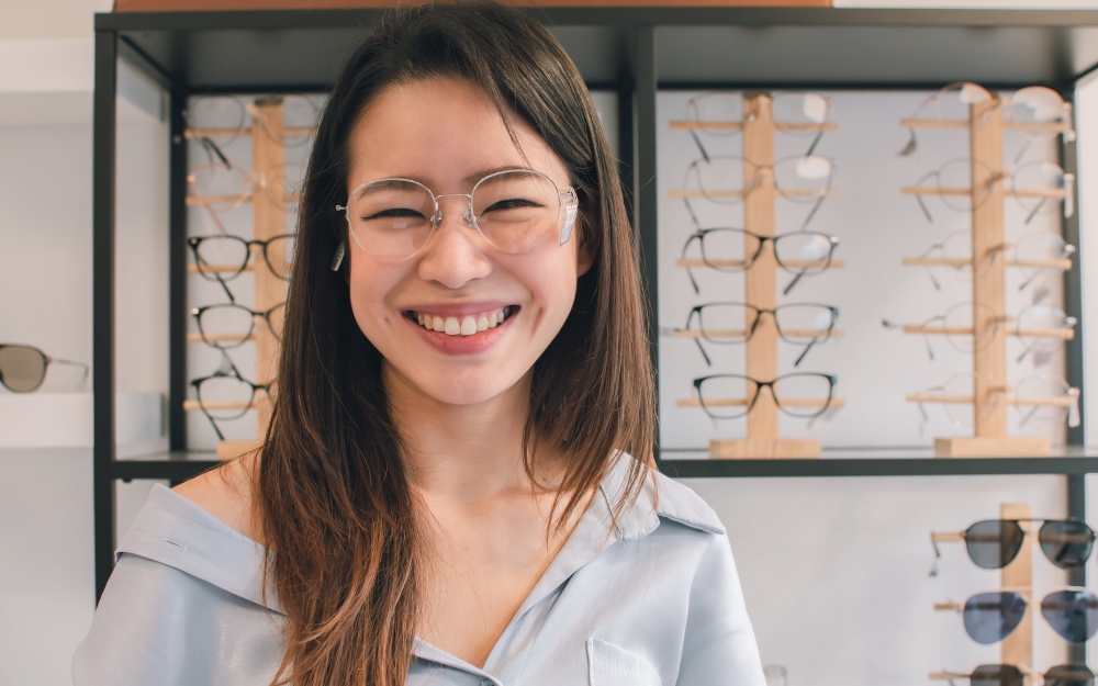 Asian beautiful young woman smiling, wearing and trying eyesight glasses in store. Sales, Discount and Optical Concept.