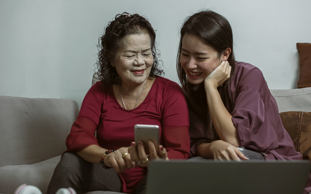 Asian old and young women online chatting by using mobile phone together in living room at home. Lifestyle, Aging, Technology and New Normal Concept.