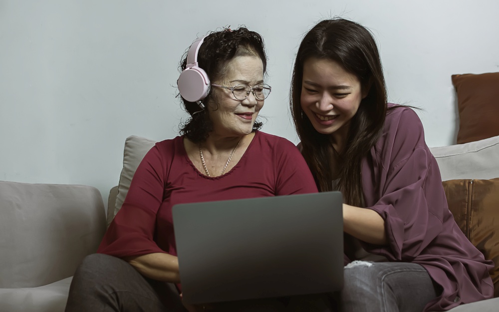 Asian old and young women online chatting by using laptop together in living room at home. Lifestyle, Aging, Technology and New Normal Concept.