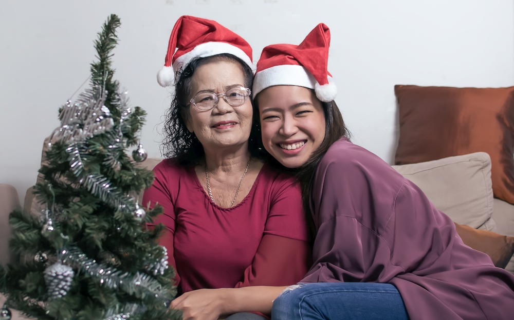 Two 60s old mother and daughter wearing santa hat, hug and sitting in living room at home with blur foreground of tree and bottle. Christmas or New Year Celebration Concept.