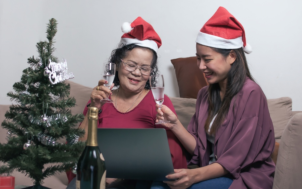 Two 60s old mother and daughter wearing santa hat, drinking and online chat with other people by using laptop while sitting in living room at home with blur foreground of tree and bottle. Christmas or New Year Celebration Concept.