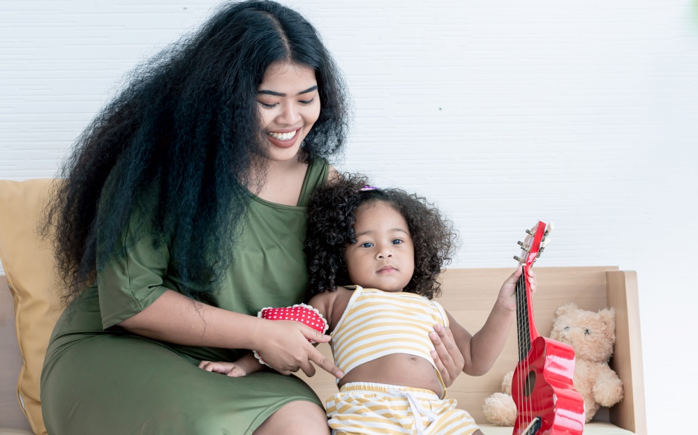 African black mother with afro hair playing ukulele with her little daughter in living room at home. Education and Family Concept.