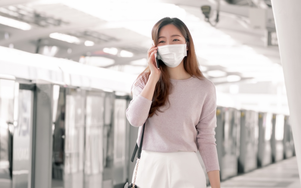 Asian woman wearing face mask and dressing casual business talking on mobile phone while standing on railway station and going to work. New Normal, Socian Distancing and Transportation Concept.