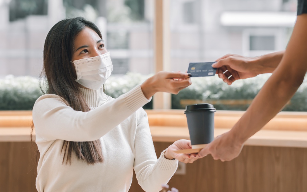 Asian beautiful woman wearing mask to protect virus and paying for coffee by credit card. Lifestyle, New Normal and Finance Concept.
