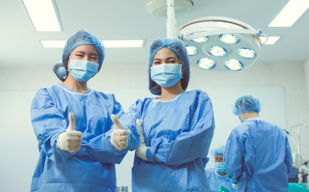 Portrait of two asian female doctors making thump up with blur background of medical team doing surgical operation at hospital