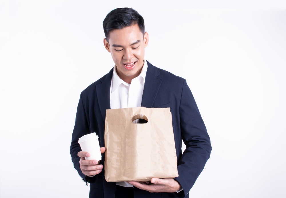 Asian handsome business man smiling, holding cup of coffee and paper bag for food delivery. He wearing formal suit and standing on white background.