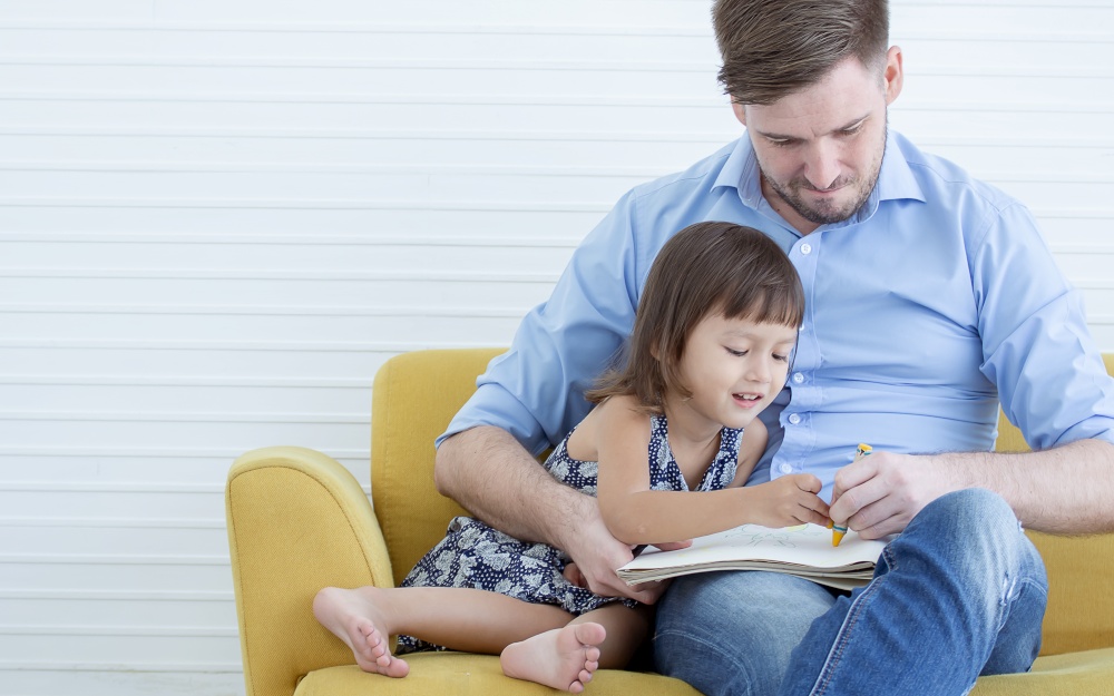 A caucasian handsome father is drawing together with his daughter while sitting on sofa in living room
