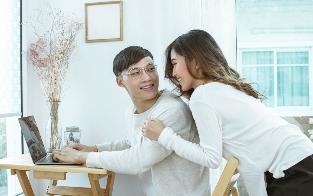 Asian lovely couple smiling and looking at each other while using laptop and working at home. Lifestyle, Valentine and Love Concept.