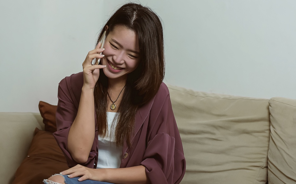 Asian beautiful woman smiling and talking on her mobile phone while sitting on sofa in living room at home. Lifestyle and Technology Concept.