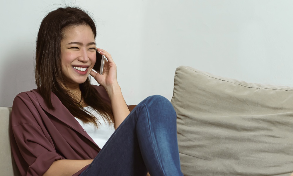 Asian beautiful woman smiling and talking on her mobile phone while sitting on sofa in living room at home. Lifestyle and Technology Concept.