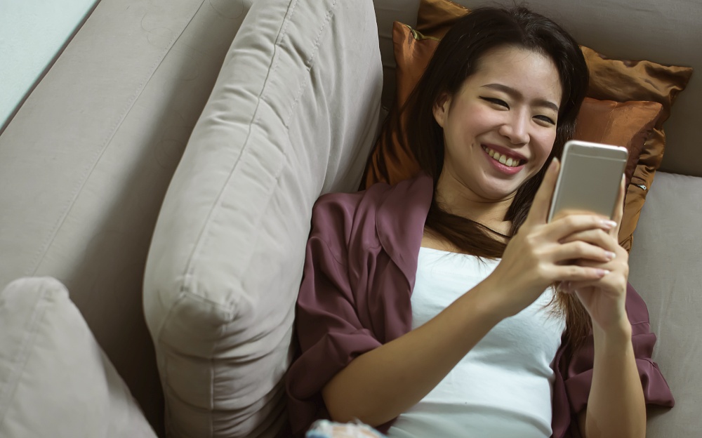Asian young beautiful woman smiling and chatting by using mobile phone  while relaxing in living room at home. Lifestyle, Technology and New Normal Concept.
