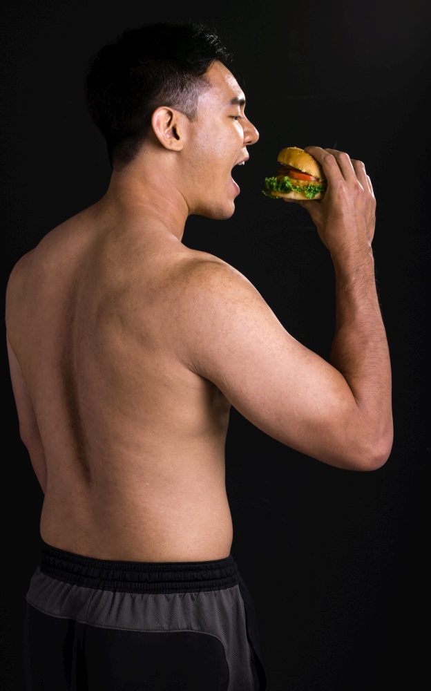 Portrait of asian young muscular man standing and eating hamburger on black background. Healthy and food Concept.