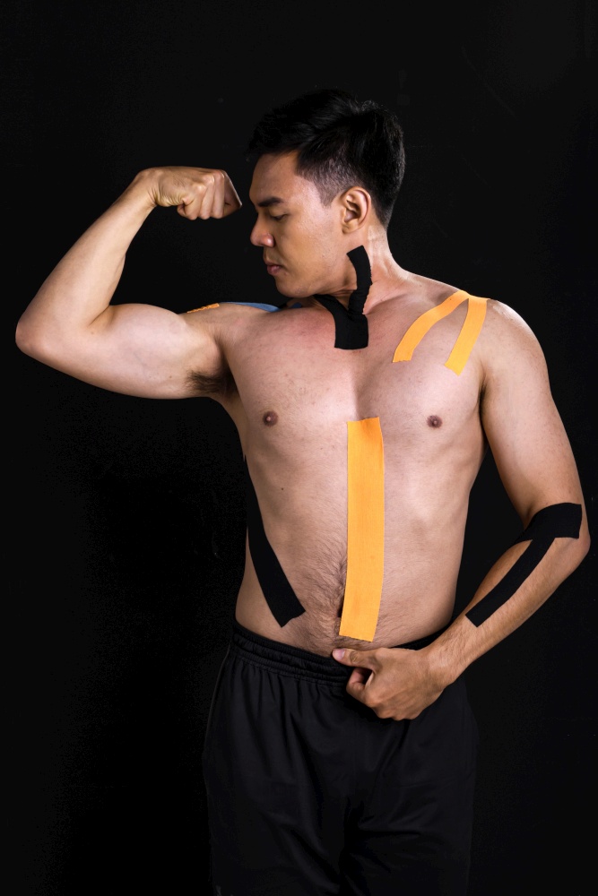 Portrait of asian young muscular man getting pain and putting kinesiology tape on his body while standing on black background with copy space. Healthy and Medical Concept.