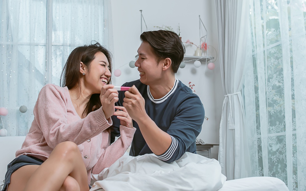 An asian lovely couple is very happy with pregnancy test while sitting on bed at home. Lifestyle Concept.