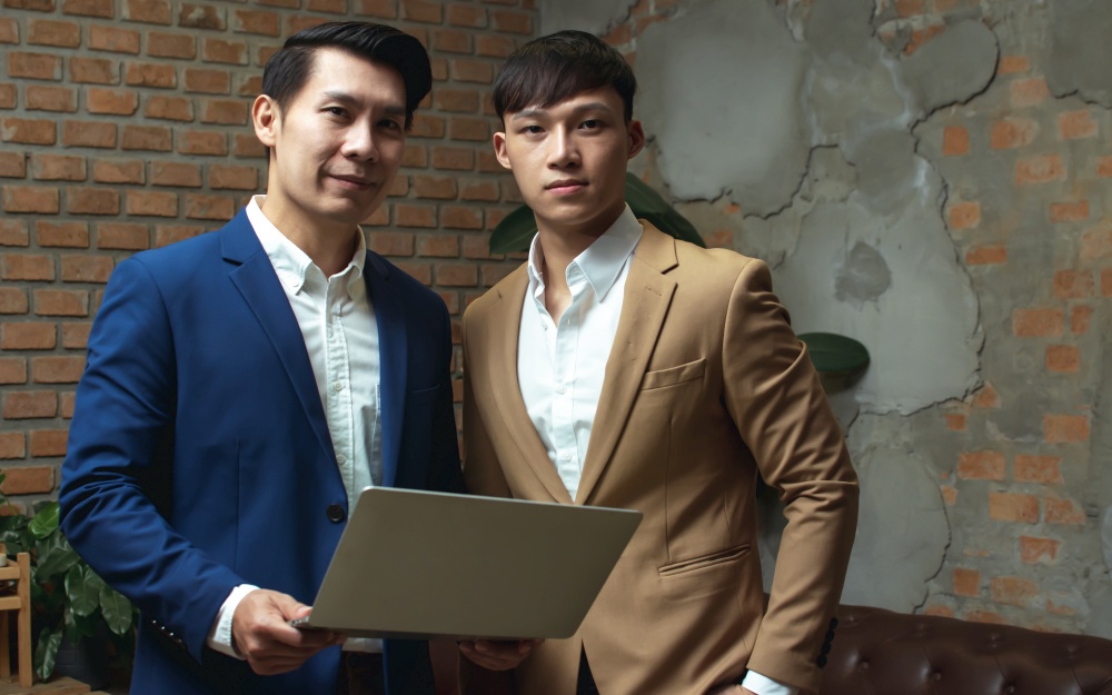 Portrait of two asian handsome business men standing, wearing formal trendy suit and holding laptop