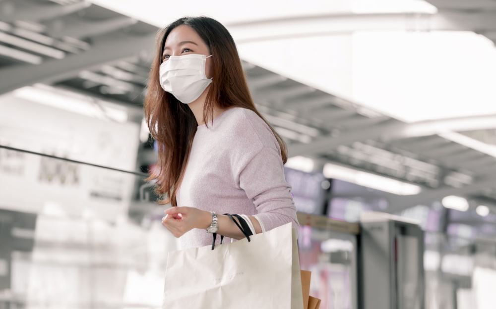 Asian woman wearing mask to protect virus, holding shopping bags and waiting for train on platform station. Sales Discount, Lifestyle and New Normal Concept.