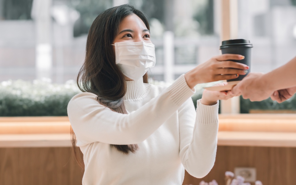 Asian woman wearing mask to protect virus and pick up coffee served by a waiter. New Normal and Social Distancing Concept.