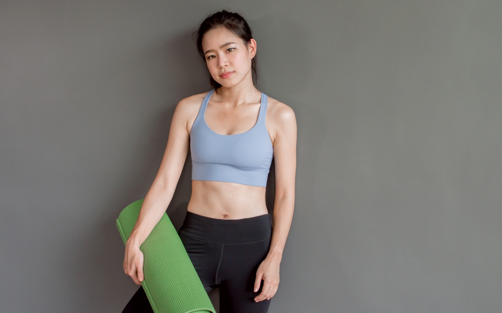 Portrait of Asian beautiful healthy young woman wearing sport bra, holding yoga mat and standing  in gym