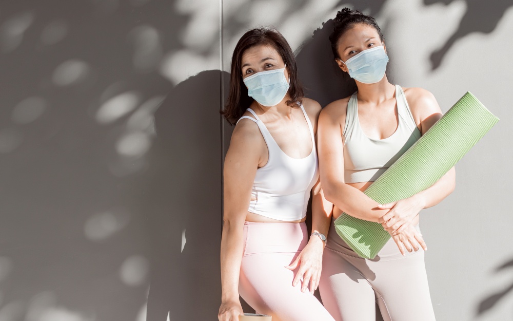 Two asian women wearing sport bra, mask to protect virus while holding yoga mat and standing  in gym. Social Distancing and Sport concept.