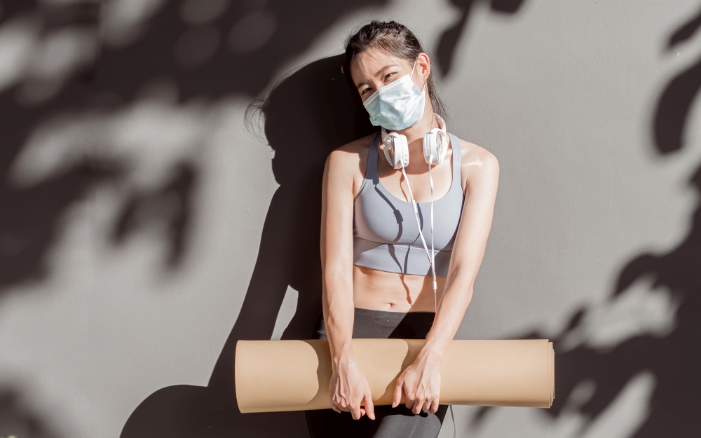 Asian woman wearing sport bra, mask to protect virus while holding yoga mat and standing  in gym. Social Distancing and Sport concept.