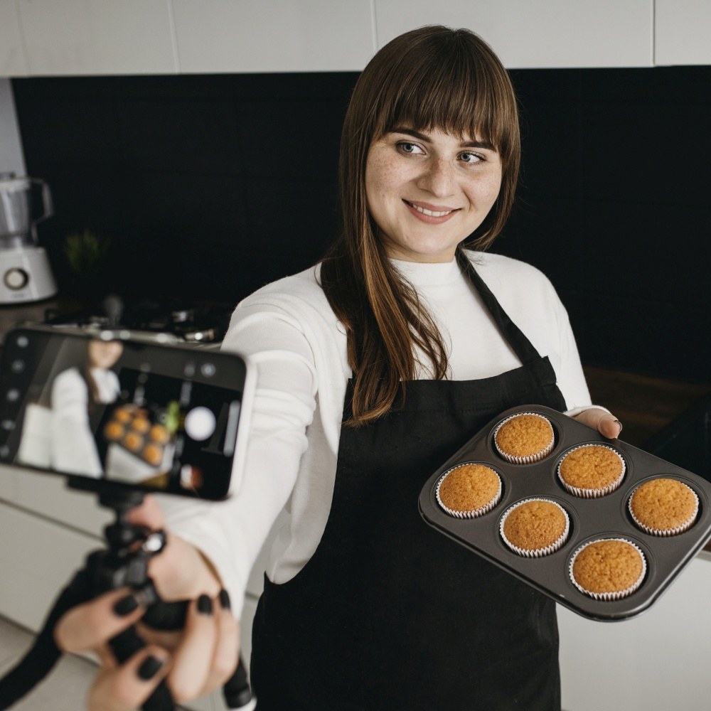smiley female blogger recording herself with smartphone while preparing muffins