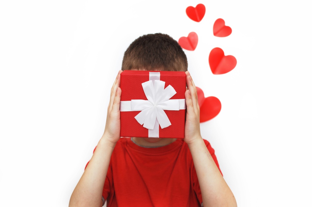 a young man holding a small present box with red hearts on a white background. child covers his face with a box. a young man holding a present box with red hearts on a white background. child covers his face with a box