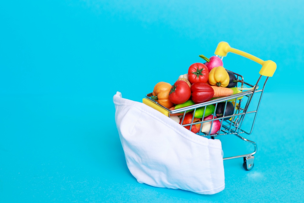 Shopping cart with protective medical mask with fruits and vegetables on a blue background. Safe and online shopping on quarantine concept.. Shopping cart with medical mask with fruits and vegetables on a blue background. online shopping on quarantine concept.