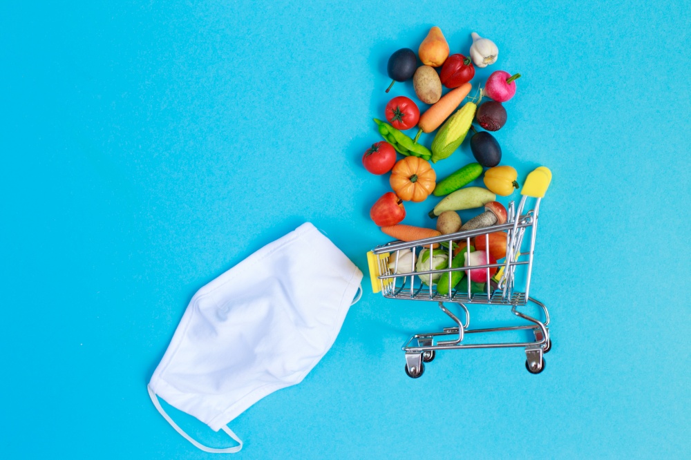 Shopping cart with protective medical mask with fruits and vegetables on a blue background. Safe and online shopping on quarantine concept.. Shopping cart with medical mask with fruits and vegetables on a blue background. online shopping on quarantine concept.