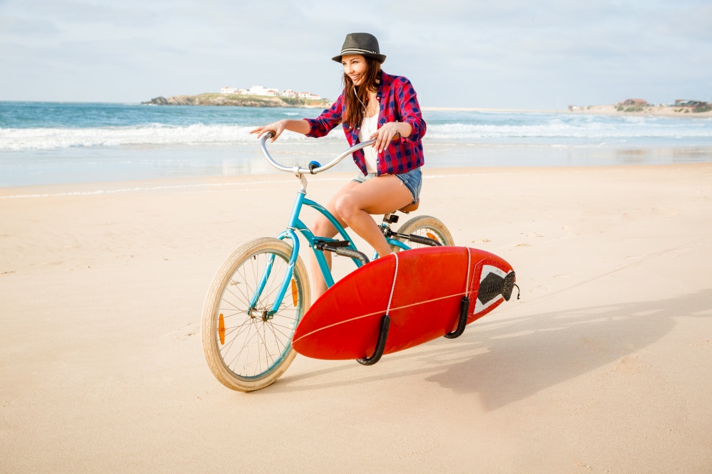 Beautiful surfer girl riding her bicycle on the beach with a surfboard