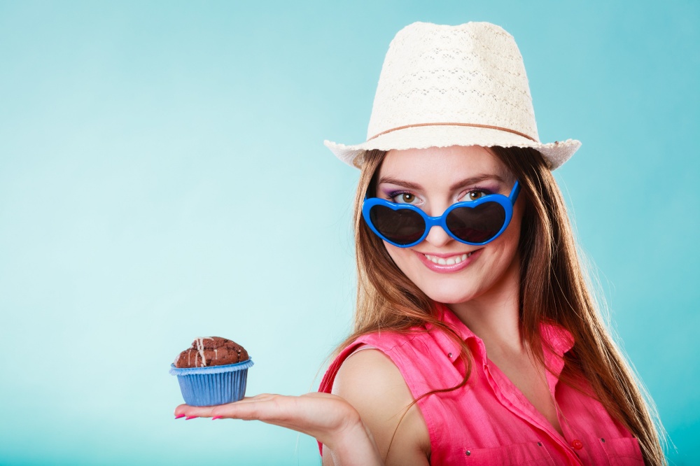 sweet food happiness and people concept. Smiling summer fashionable woman wearing straw hat heart shaped sunglasses holds cake cupcake in hand blue background