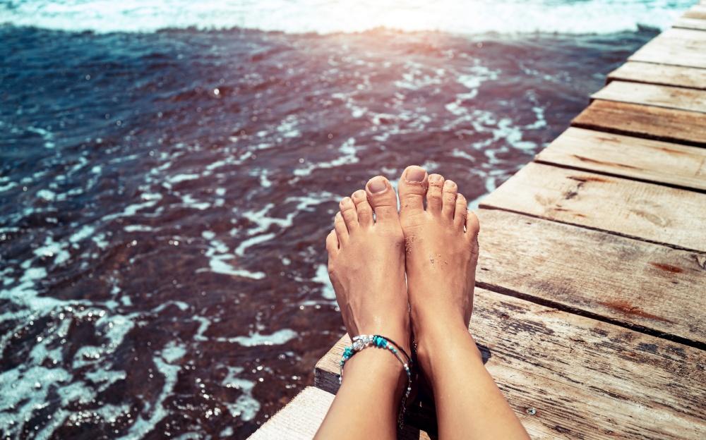 Closeup photo of a women&rsquo;s legs on the wooden pier over the sea, female with pleasure tanning on the beach, summer vacation concept