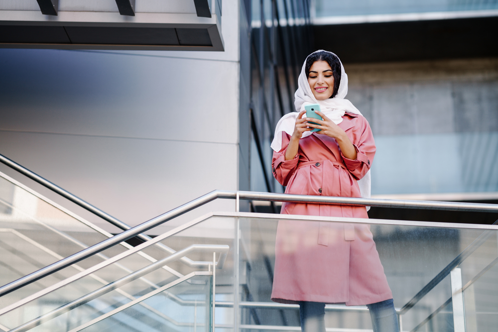 Young Arab woman wearing hijab headscarf texting message with her smartphone in a office building. Business background.. Young Muslim woman wearing hijab texting message with her smartphone.