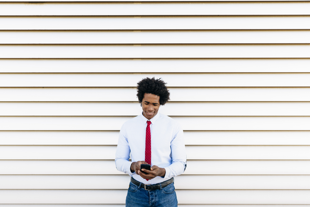 Black Businessman using a smartphone with a white blinds background. Man with afro hair.. Black Businessman using a smartphone with a white blinds background