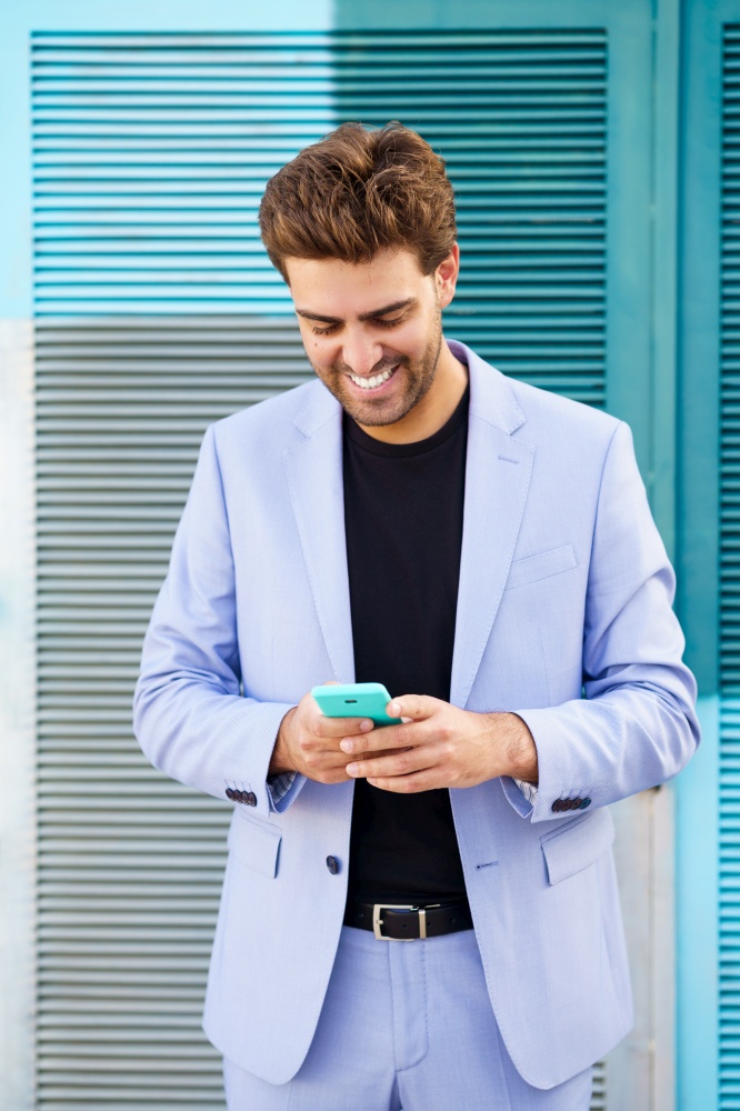 Smiling man wearing blue suit texting with a smartphone in urban background.. Smiling man wearing blue suit texting with a smartphone outdoors.