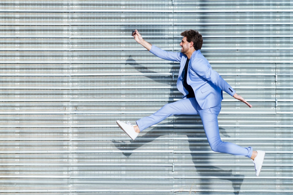 Young man wearing a suit makes a selfie with a smartphone while jumping outdoors. Man wearing a suit makes a selfie with a smartphone while jumping