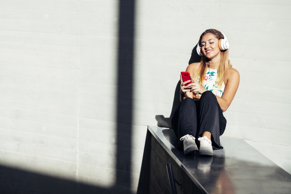 Young woman using a smartphone wearing casual clothes and headphones. Girl sitting outdoors.. Girl using a smartphone wearing casual clothes