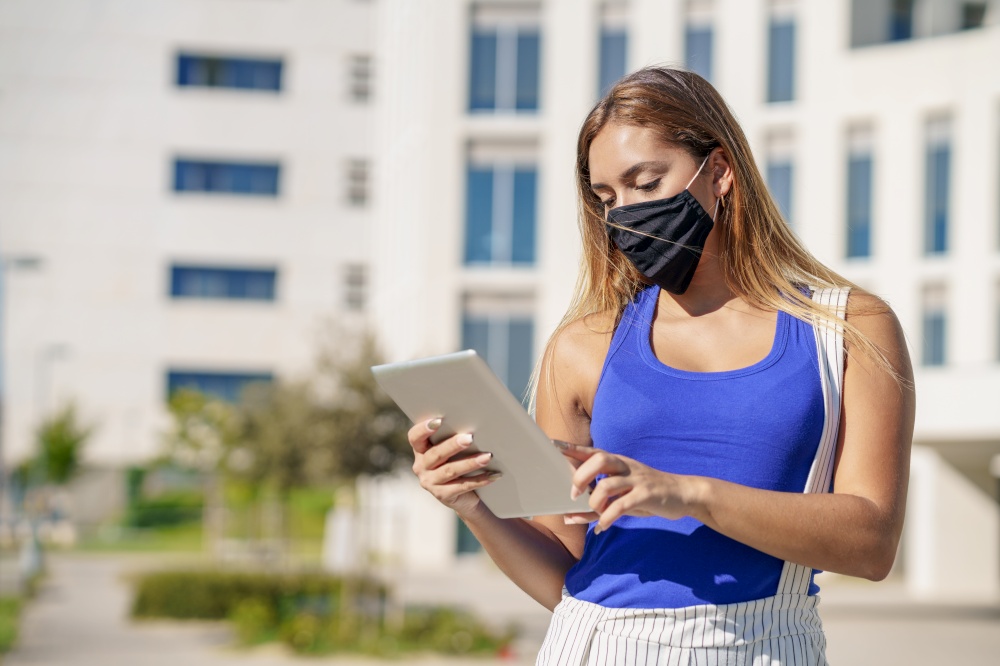 Young woman wearing a mask uses digital tablet near an office building. Young woman wearing a mask uses digital tablet outdoors.